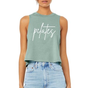 May include: A sage green sleeveless crop top featuring the word pilates written in white cursive script across the chest. The garment has a crew neckline and a relaxed, boxy fit with raw-edge armholes, paired with light blue denim jeans.