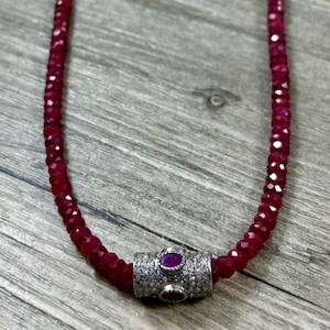 May include: A necklace featuring a strand of faceted, deep red beads. The necklace has a silver clasp with a central ruby and is displayed on a wooden surface. The beads are approximately 0.2 inches in diameter.