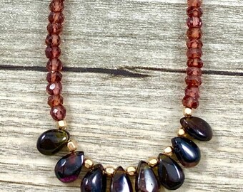 May include: A necklace made of small, faceted garnet beads with a teardrop-shaped pendant made of a larger, faceted garnet. The necklace is displayed on a purple amethyst cluster.