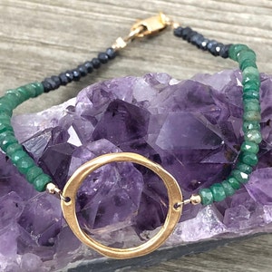 Könnte beinhalten: Ein goldener Kreisanhänger hängt an einem Armband aus grünen und schwarzen Perlen. Das Armband liegt auf einem violetten Amethystkristall.