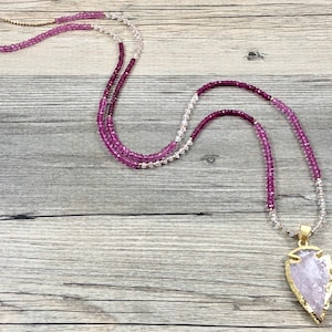 May include: A layered necklace with pink and white beads and a large, gold-trimmed, arrowhead-shaped pendant made of clear quartz.