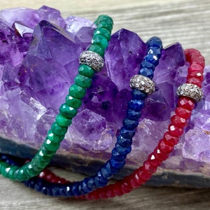 May include: Three beaded bracelets with silver charms on a purple amethyst crystal background. The bracelets are green, blue, and red.