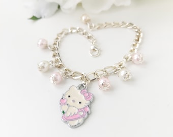 May include: A silver-toned chain bracelet featuring a central enamel charm of a white cat character wearing a pink dress and bow. The bracelet is accented with multiple dangling round beads in shades of white and soft pink, finished with a lobster clasp.