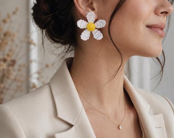 May include: A pair of stud earrings featuring a crocheted daisy design. Each earring consists of six white petals surrounding a textured yellow center. The floral motif is crafted from thread, creating a distinct, layered appearance on the earlobe.