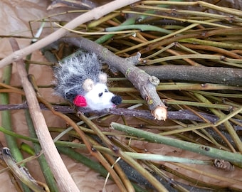 May include: A small, gray and white felt hedgehog with red accents is nestled among a pile of brown and green twigs.