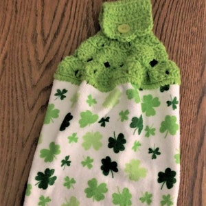 St Patrick’s hanging kitchen towel with a crocheted top.  It has shamrocks in different colors of green on white background.