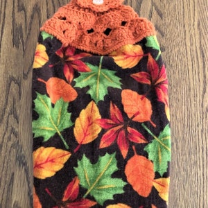 May include: A brown kitchen towel with a colorful autumn leaf print and a crocheted orange top with a button.