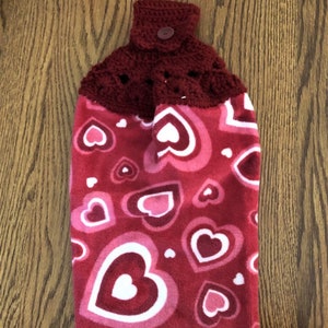 May include: A red and pink heart-patterned kitchen towel with a crocheted top and button closure. The towel features various sizes of hearts in shades of red, pink, and white. The top is a dark burgundy color.