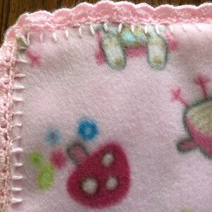 May include: Pink fleece baby blanket with a crocheted pink border. The blanket features a variety of colorful animal and floral designs.