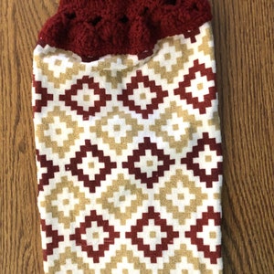May include: A red and gold kitchen towel with a crocheted top and a button. The towel has a geometric pattern.