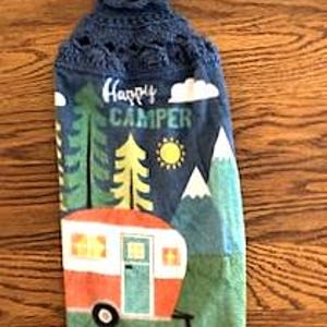 May include: A blue and green hanging kitchen towel with a crochet top. The towel features a camper, trees, mountains, a sun, and the words "Happy Camper". The camper is orange and white, and the trees are green and yellow.