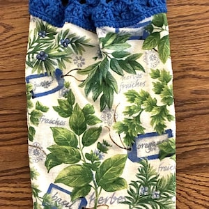 May include: A hanging kitchen towel featuring a white background with a green herb pattern, including illustrations of basil, oregano, and juniper, with the text "herbes fraiches". The top has a blue crocheted topper with a matching blue button.
