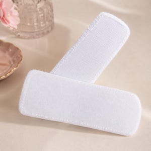 May include: Two white, rectangular, fabric pads with rounded corners are displayed at an angle. One pad is positioned over the other, with a portion of the top pad visible. The pads have a soft, textured appearance, suggesting a cleaning or cosmetic product.
