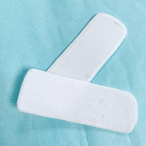 May include: Two white, rectangular fabric patches with rounded corners are shown against a light blue background. One patch is positioned over the other, creating a layered effect. The patches have a soft, textured appearance.