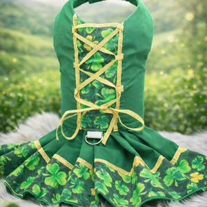 May include: Green dog dress with a halter top and a skirt. The dress features gold trim and a shamrock pattern. The bodice has a gold criss-cross design. A metal ring is attached to the front of the dress.