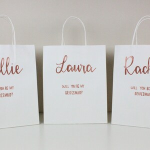 May include: Three white gift bags with rose gold lettering. The bags have the names Kallie, Laura, and Rachel printed on them, along with the question "Will you be my bridesmaid?"