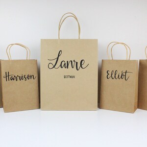 May include: Five brown paper gift bags with black lettering. The bags are labeled with the names "Conan", "Harrison", "Lanre Bestman", "Elliot", and "Nick".
