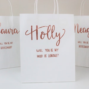 May include: Three white gift bags with rose gold lettering. The bags are personalized with the names "Laura", "Holly", and "Meaghan". Each bag has a message asking the recipient to be a bridesmaid or maid of honor.