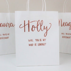 May include: Three white gift bags with rose gold lettering. The bags have the names "Laura", "Holly", and "Meaghan" written on them. The bags also have the text "Will you be my bridesmaid?" and "Will you be my maid of honour?" written on them.