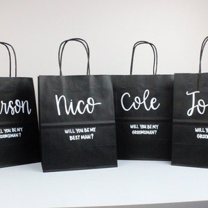 May include: Four black paper gift bags with white lettering. Each bag has a name printed on the front: Karson, Nico, Cole, and Josh. Each bag also has the text "Will you be my groomsman?" or "Will you be my best man?"