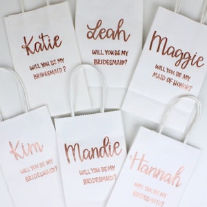 May include: Six white gift bags with rose gold lettering. Each bag has a different name and a message asking "Will you be my bridesmaid?" or "Will you be my maid of honor?"