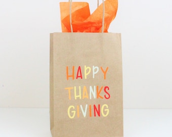 May include: A brown paper gift bag with a bright orange tissue paper peeking out of the top. The bag has the words "HAPPY THANKSGIVING" written on it in colorful letters.