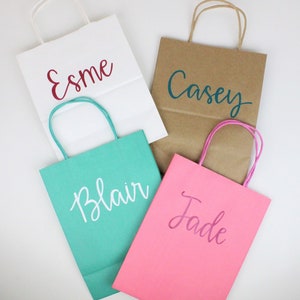 Pu&ograve; includere: Quattro sacchetti regalo con manici, ognuno con un nome diverso scritto sul davanti. I sacchetti sono bianchi, marroni, turchese e rosa. I nomi sono Esme, Casey, Blair e Jade.