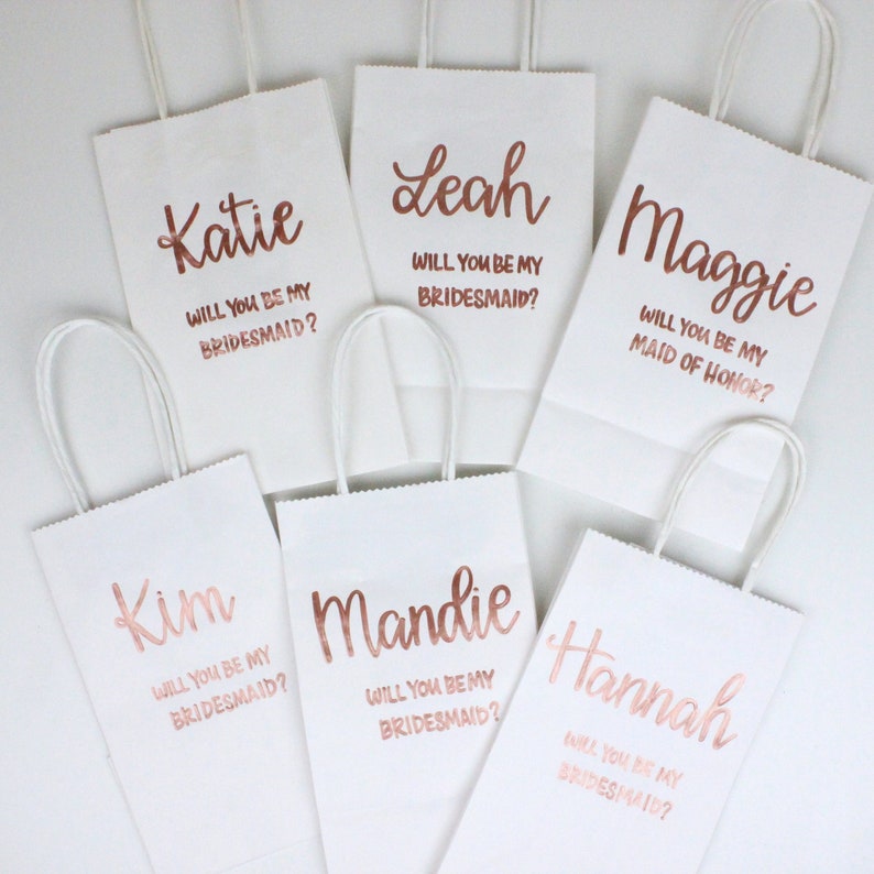May include: Six white gift bags with rose gold lettering. Each bag has a different name and a message asking "Will you be my bridesmaid?" or "Will you be my maid of honor?"