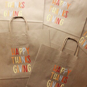 Puede incluir: Se muestran varias bolsas de la compra de papel marr&oacute;n con asas. Cada bolsa tiene las palabras "HAPPY THANKS GIVING" impresas en letras coloridas. Las bolsas son adecuadas para regalos o recuerdos de fiesta.