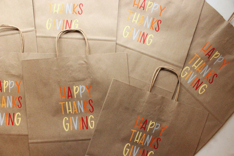 Puede incluir: Se muestran varias bolsas de la compra de papel marr&oacute;n con asas. Cada bolsa tiene las palabras "HAPPY THANKS GIVING" impresas en letras coloridas. Las bolsas son adecuadas para regalos o recuerdos de fiesta.