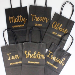May include: Six black paper gift bags with gold lettering. The bags are labeled with the names "Matty", "Trevor", "Albie", "Ian", "Sheldon", and "Isaiah". The bags are for a wedding party and include the titles "Groom", "Best Man", and "Groomsman".