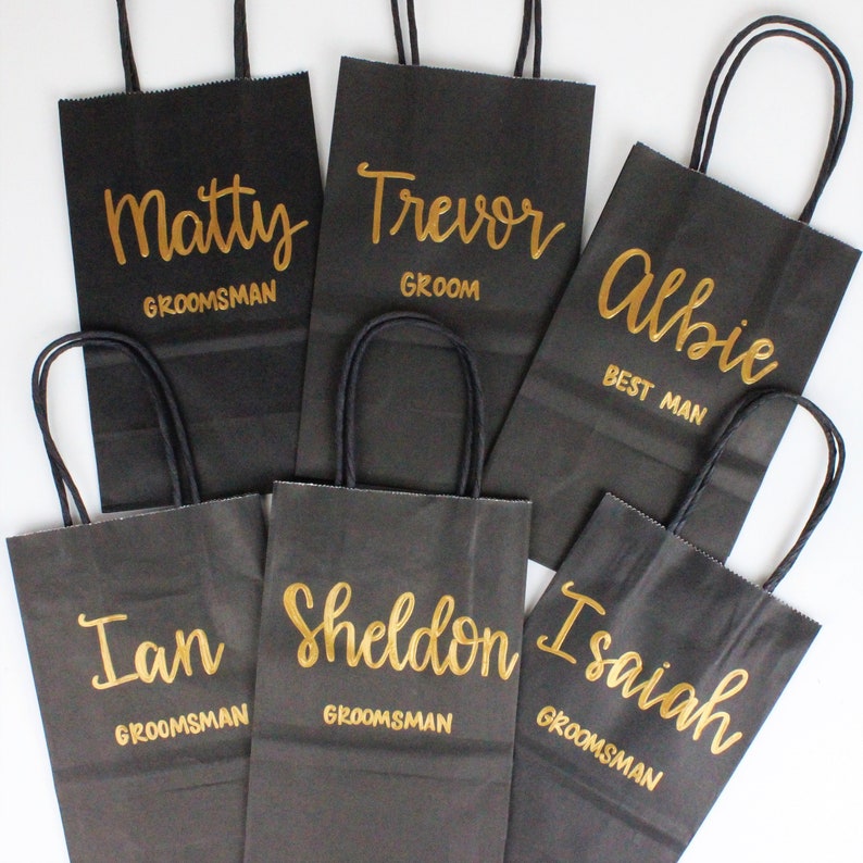 May include: Six black paper gift bags with gold lettering. The bags are labeled with the names "Matty", "Trevor", "Albie", "Ian", "Sheldon", and "Isaiah". The bags are for a wedding party and include the titles "Groom", "Best Man", and "Groomsman".