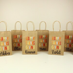 Puede incluir: Ocho bolsas de regalo de papel marr&oacute;n con asas de papel retorcido. Cada bolsa presenta las palabras "HAPPY THANKSGIVING" en letras coloridas, con un nombre impreso debajo. Los nombres son Zach, Abby, Ollie, Kimmie, Marc, Violet, Ellie y Celine.