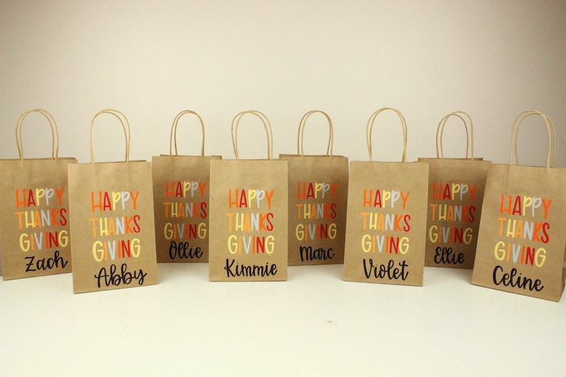 Puede incluir: Ocho bolsas de regalo de papel marr&oacute;n con asas de papel retorcido. Cada bolsa presenta las palabras "HAPPY THANKSGIVING" en letras coloridas, con un nombre impreso debajo. Los nombres son Zach, Abby, Ollie, Kimmie, Marc, Violet, Ellie y Celine.