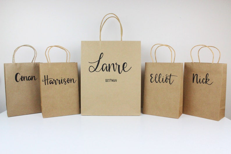 May include: Five brown paper gift bags with black lettering. The bags are labeled with the names "Conan", "Harrison", "Lanre Bestman", "Elliot", and "Nick".