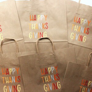 Puede incluir: Bolsas de papel marr&oacute;n con las palabras "HAPPY THANKS GIVING" impresas en letras coloridas. Las bolsas tienen asas de papel retorcido y son adecuadas para regalos o recuerdos de fiesta.