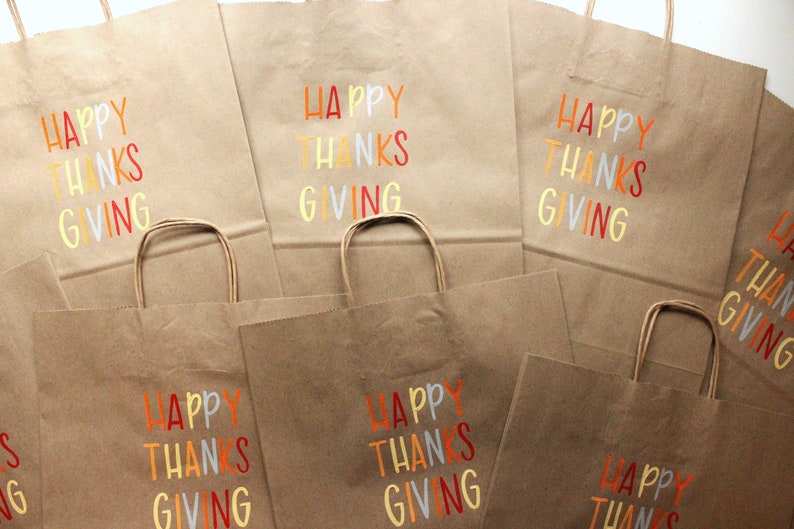Puede incluir: Bolsas de papel marr&oacute;n con las palabras "HAPPY THANKS GIVING" impresas en letras coloridas. Las bolsas tienen asas de papel retorcido y son adecuadas para regalos o recuerdos de fiesta.