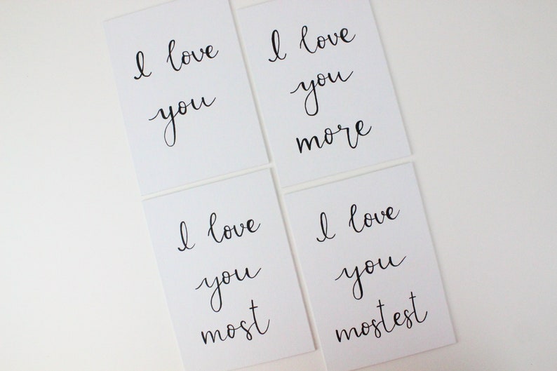 Puede incluir: Cuatro tarjetas blancas con caligraf&iacute;a negra que dice "Te amo", "Te amo m&aacute;s", "Te amo m&aacute;s", y "Te amo m&aacute;s que nada".