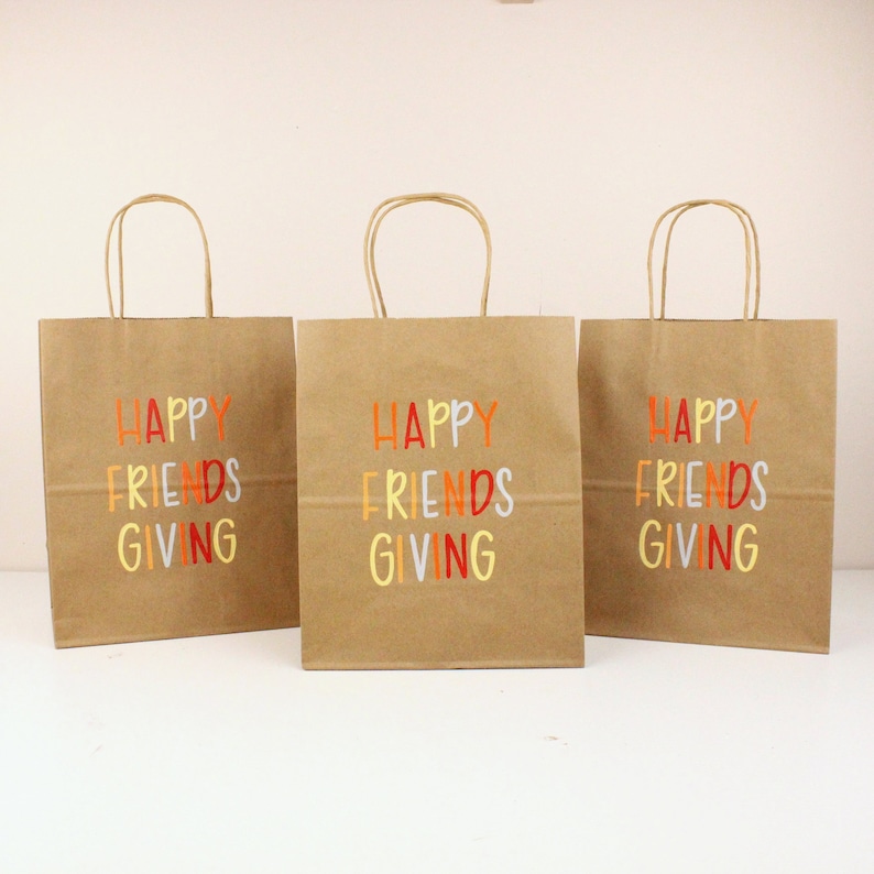 Puede incluir: Tres bolsas de regalo de papel marr&oacute;n con asas de papel retorcido. Cada bolsa tiene las palabras "HAPPY FRIENDS GIVING" impresas en letras coloridas. Las bolsas est&aacute;n dispuestas en fila sobre un fondo blanco.