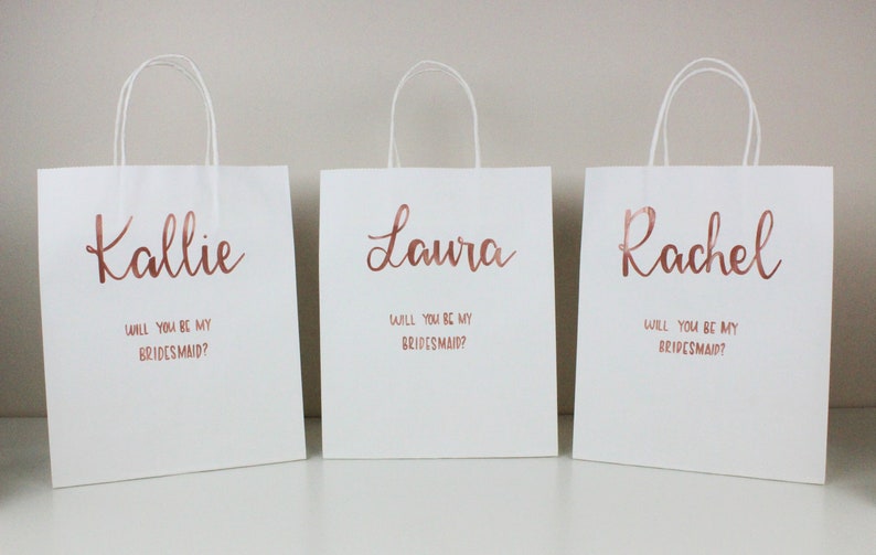 May include: Three white gift bags with rose gold lettering. The bags have the names Kallie, Laura, and Rachel printed on them, along with the question "Will you be my bridesmaid?"