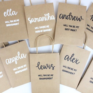 May include: A collection of brown paper gift bags with white and black lettering. The bags are personalized with names and the text "Will you be my bridesmaid?" or "Will you be my groomsman?"