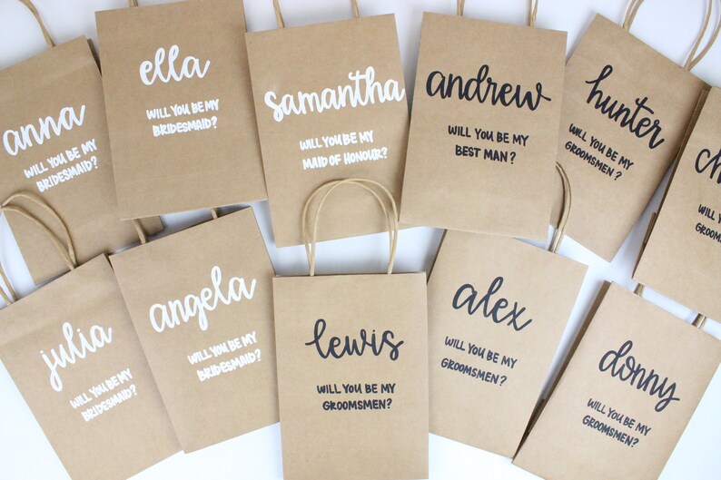 May include: A collection of brown paper gift bags with white and black lettering. The bags are personalized with names and the text "Will you be my bridesmaid?" or "Will you be my groomsman?"