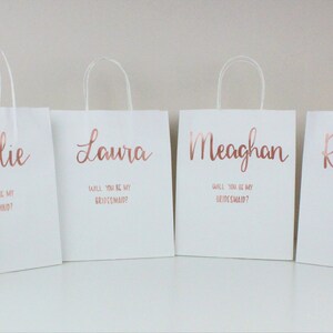 May include: Six white gift bags with rose gold lettering. Each bag has a different name on it: Holly, Kallie, Laura, Meaghan, Rachel, and Olivia. The text on the bags reads "Will you be my maid of honor?" or "Will you be my bridesmaid?"