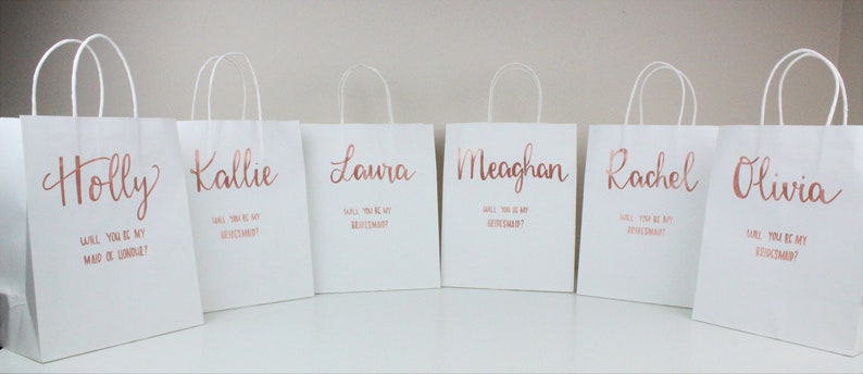 May include: Six white gift bags with rose gold lettering. Each bag has a different name on it: Holly, Kallie, Laura, Meaghan, Rachel, and Olivia. The text on the bags reads "Will you be my maid of honor?" or "Will you be my bridesmaid?"