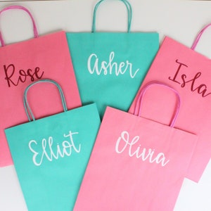 Pu&ograve; includere: Cinque sacchetti regalo di carta colorati con manici. I sacchetti sono rosa e turchese e hanno nomi scritti in lettere bianche: Rose, Asher, Isla, Elliot e Olivia.