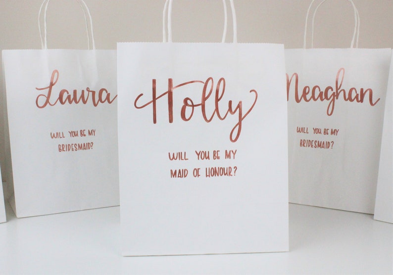 May include: Three white gift bags with rose gold lettering. The bags have the names "Laura", "Holly", and "Meaghan" written on them. The bags also have the text "Will you be my bridesmaid?" and "Will you be my maid of honour?" written on them.