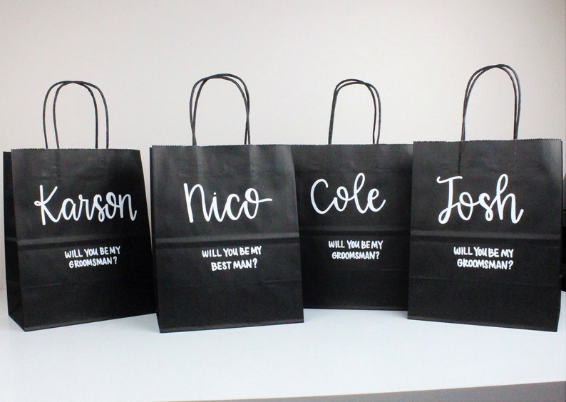 May include: Four black paper gift bags with white lettering. Each bag has a name printed on the front: Karson, Nico, Cole, and Josh. Each bag also has the text "Will you be my groomsman?" or "Will you be my best man?"