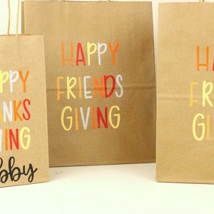 Puede incluir: Tres bolsas de regalo de papel marr&oacute;n con texto colorido. Las bolsas dicen "Happy Thanks Giving" y "Happy Friends Giving". Una bolsa est&aacute; personalizada con el nombre "Abby". El texto es en naranja, rojo, amarillo y blanco.