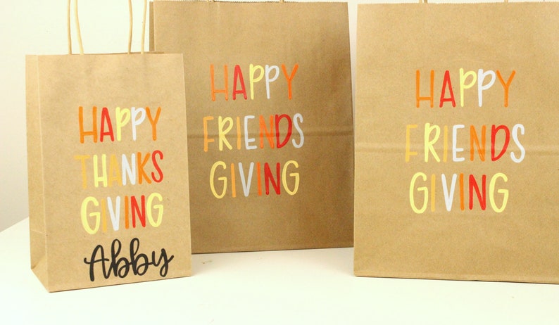 Puede incluir: Tres bolsas de regalo de papel marr&oacute;n con texto colorido. Las bolsas dicen "Happy Thanks Giving" y "Happy Friends Giving". Una bolsa est&aacute; personalizada con el nombre "Abby". El texto es en naranja, rojo, amarillo y blanco.