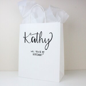 May include: White gift bag with black text that says "Kathy" and "Will you be my bridesmaid?"